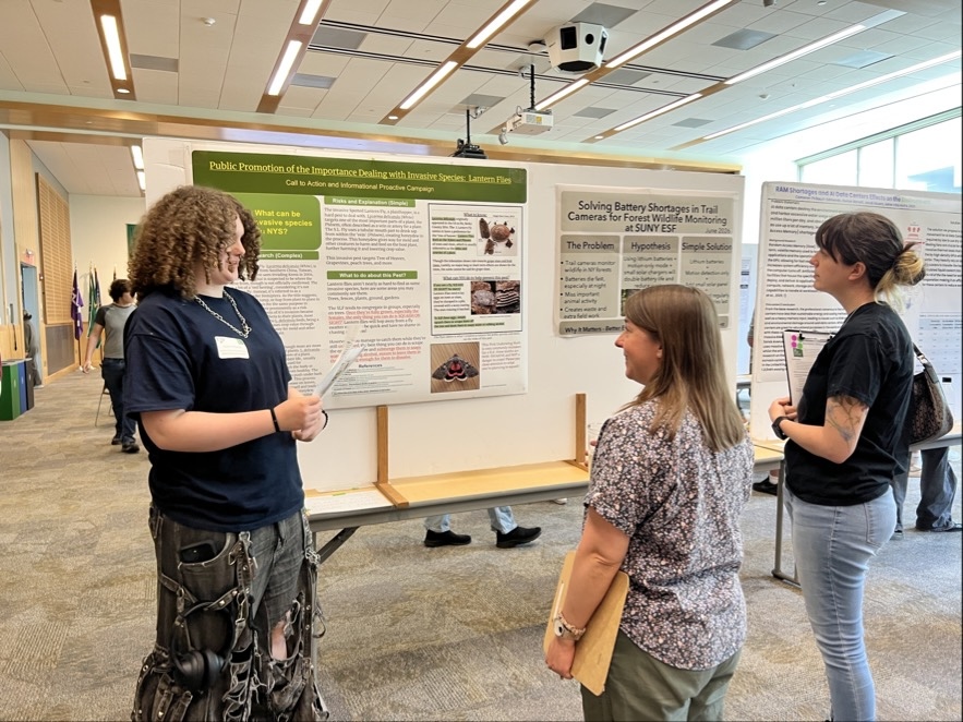 A presenter stands beside a research poster explaining the project to two attendees in a bright indoor event space. Several large display boards with detailed text, images, and headings are arranged throughout the room.