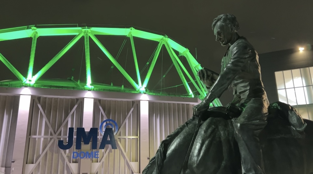 A nighttime photo shows a brightly lit green JMA Wireless Dome behind a bronze statue of Abraham Lincoln reading a book while riding a horse. The dome’s exterior beams glow vividly against the dark sky, while the statue is prominently featured in the foreground. 