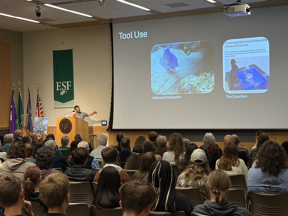 A person stands at a podium in a packed auditorium, presenting a slide titled “Tool Use” that features examples of animal tool use from National Geographic and The Guardian. Audience members fill the seats as the presentation is projected on a large screen beside an ESF banner.