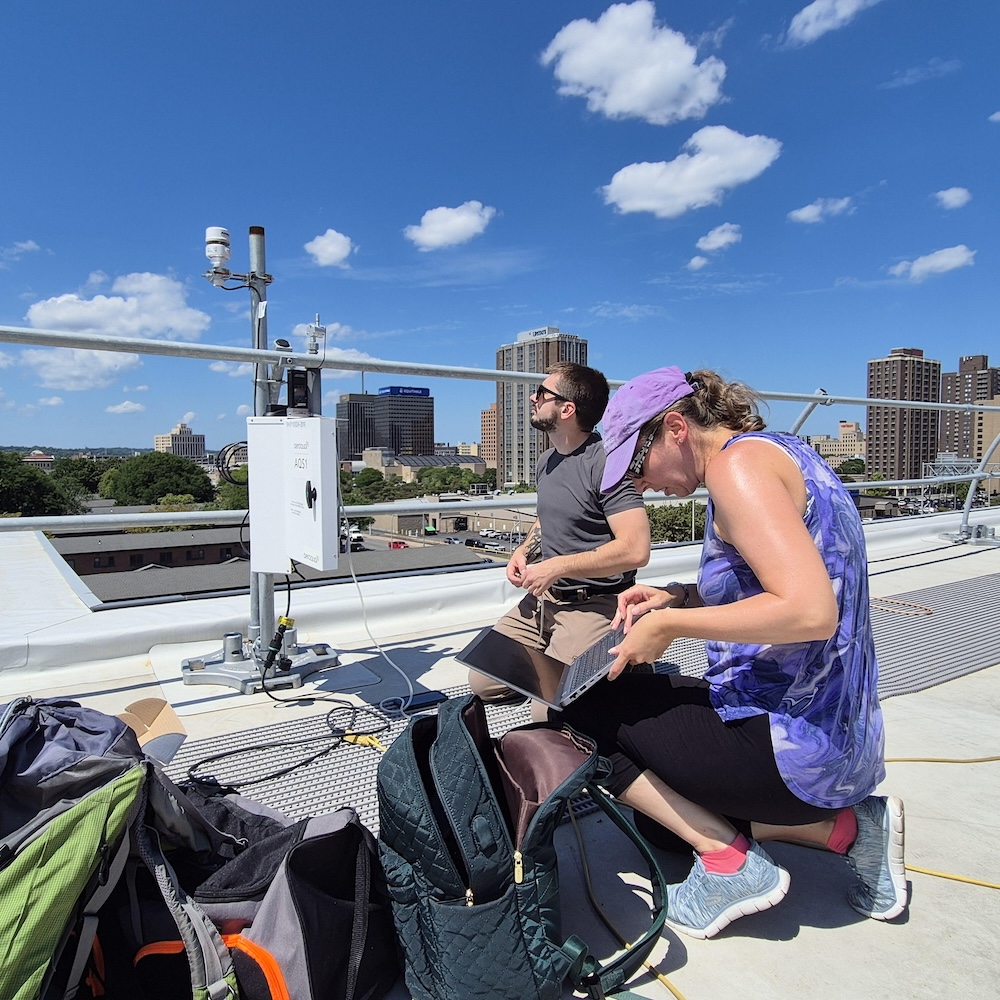 Two people work with monitoring equipment on a rooftop overlooking a city skyline on a bright day with scattered clouds. One person kneels beside an open laptop connected to air-quality monitoring instruments, while another stands nearby next to the mounted sensors and data collection equipment.