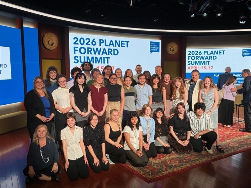 A large group of people pose together on a stage in front of two large screens reading “2026 Planet Forward Summit,” with additional text showing the dates April 15–17. The group is arranged in rows on a patterned rug under stage lighting.