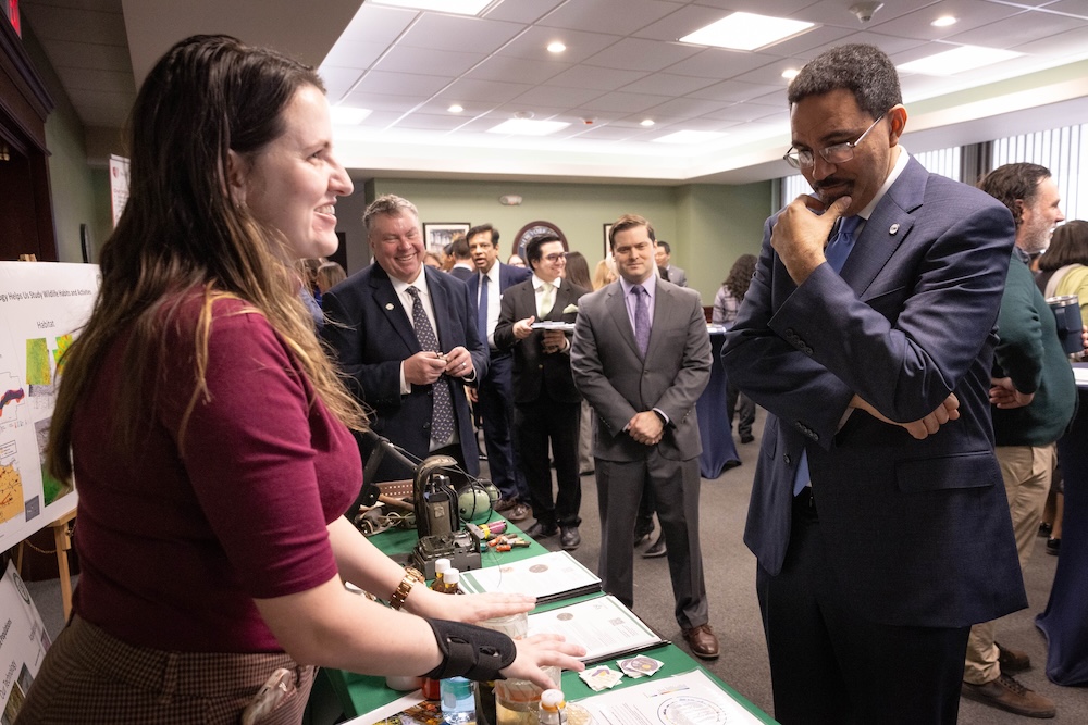 A person stands behind a table displaying project materials while speaking to another person across from the table. Several other people stand in the background observing the interaction.