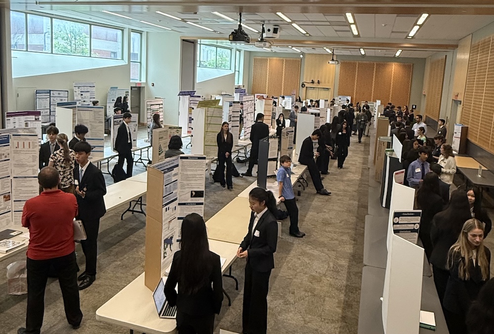 A large group of students stand beside poster boards arranged in rows inside a bright hall, presenting research projects on tables. Attendees move throughout the room, stopping to view displays and speak with presenters.
