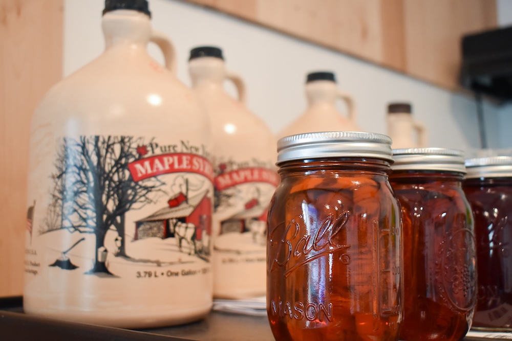 Several glass jars filled with amber maple syrup sit on a shelf in the foreground. Behind them, large cream‑colored jugs labeled “Pure New York Maple Syrup” are slightly out of focus.