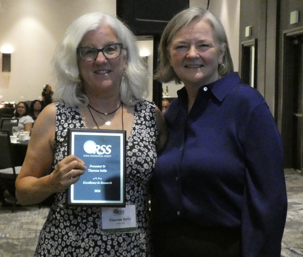 Two people stand together indoors at an awards event while one person holds a plaque recognizing excellence in research. The plaque displays the CRSS (Rural Sociological Society) logo and the year 2026.