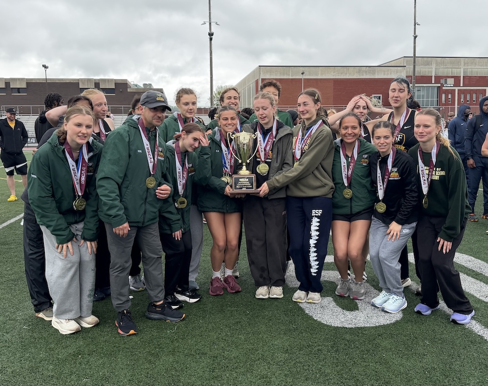 A group of student athletes stands closely together on an outdoor track field, wearing medals around their necks and holding a large trophy at the center of the group. Campus buildings and other people are visible in the background under an overcast sky.
