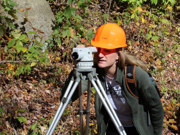 Student in an orange hard hat uses surveying equipment in a wooded setting.