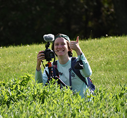 Lilly Kramer holding a camera in one hand and thumbs up in another standing in a field