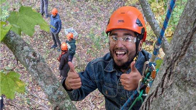 Student in a hard hat giving a thumbs up in a tree with students below.