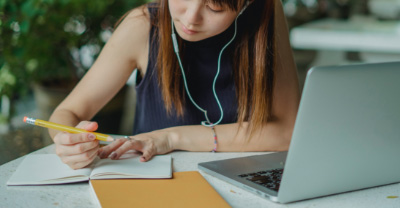 Person taking notes at a desk in front of a laptop computer.