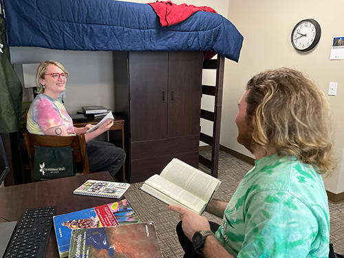 Two students in Centennial Triples room. A student has a book open in their hand and anotehr student is siting on a chair by the bunk bed