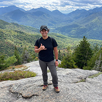 alexis rocio guillermo standing on rocks wearing a black tshirt and grey pants. The background show mountain ranges