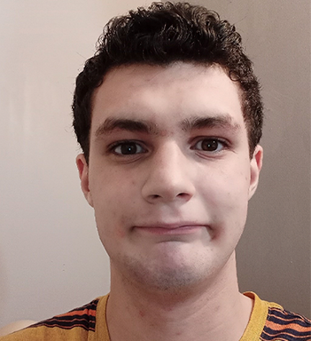 Emeric Panyky smiles at the camera with his mouth closed. He has brown, curly  hair that is kept short. He is wearing yellow t-shirt with orange and black stripes