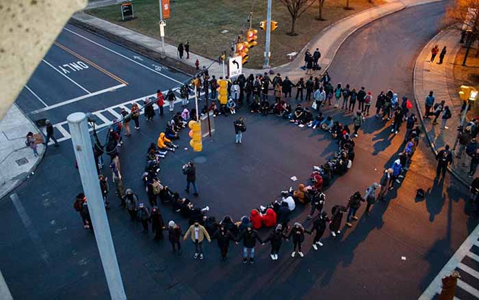 A large group of students and community members create two circles with their bodies - one within another. The students in the outer circle are holding hands. They are gathering together in an intersection on Syracuse University's campus.