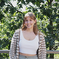 A headshot of Abbey Leibert. She is wearing a white tank top and a white-and-brown patterned sweater. She has long, blond hair. She is standing in front of trees. She is smiling with her teeth.
