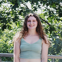 A headshot of Allie Kopinski. She is wearing a blue tank top. She has brown curly hair. She is standing in front of trees. She is smiling with her teeth.