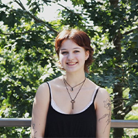 A headshot of Allison LeClair. They are wearing a black tank top. They are wearing two necklaces. Their hair is short and tucked behind their ears. They are standing in front of trees. They are smiling with their teeth.