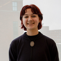 A headshot of Allison LeClair. Allison is smiling with their hands behind their back. Allison has red hair with bangs and is wearing a black sweater with a bolo tie.