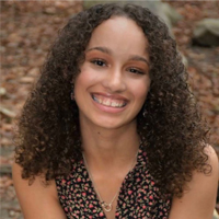 Asia Frantz smiles at the camera with teach. She wear a floral blouse and has a necklace. She has curly brown hair that extends just past her shoulders.