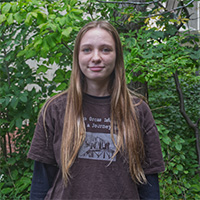 Person in a brown shirt smiles in front of a background of trees