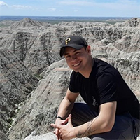 A headshot of Christopher Emerson. He is wearing a black shirt and a black hat. He is crouching at the top of a canyon. He is smiling with his teeth.