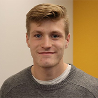 A headshot of Christopher. Christopher is wearing a grey sweater. Christopher has short brown hair. Christopher is smiling.