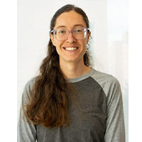 A headshot of Emma Goertzen smiling against a white background. She is wearing a gray shirt, glasses, and sports chest length hair.