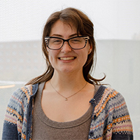 A headshot of Erin Lutz. Erin is smiling and has long brown hair with half of it pulled back. She is wearing glasses, a gray shirt, and a blue and orange colorful cardigan over top.