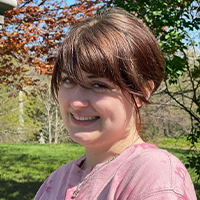 A headshot of Erin Lutz. She is wearing a pink shirt. Her hair is brown and pulled back behind her head. She is standing in a park. She is smiling with her teeth. 
