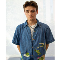 Everett Tucker with their hands behind their back against a white background. They sport short hair and a necklace. They’re wearing a blue shirt with embroidered nature prints.