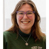 A headshot of Grace Bedford. She is wearing a green SUNY ESF crew neck and purple glasses. She has brown hair to her shoulders. She is smiling with her teeth. 