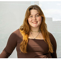Gracie Gilcrist smiling in front of a white background. She is wearing a brown shirt, stacked necklaces and chest length red hair