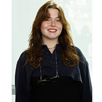 A headshot of Grace Gilcrist. She is wearing a black top and her glasses are hanging on ther neck