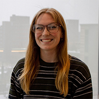 A headshot of Ivy Ven Roy. She is wearing a striped sweater and glasses. Her hair is long and strawberry blonde. She is smiling with her teeth.  