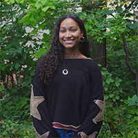Person in a black sweater with brown patches and a necklace smiles in front of a background of trees