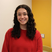 a headshot of Julia. She is wearing a red sweater. She has wavy dark brown hair. Julia is smiling with her teeth. She is inside in front of a beige and yellow wall.