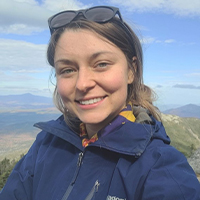 A headshot of Leah Prudent. Her hair is blond and pulled back behind her head. She is wearing a navy-blue jacket. She has sunglasses on top of her head. There is a blue sky with clouds behind her. She is smiling with her teeth.