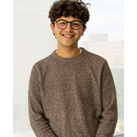 Max Loccisano wearing a brown crew neck sweater. He has curly short hair and wears glasses