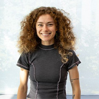 Melanie Costello smiling against a white background. She sports curly, shoulder length hair, and a black shirt with pink embroidery.