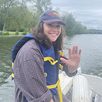 A headshot of Rebecca Rowe. She is wearing a blue baseball hat, blue life jacket, and a plaid shirt. She has dark brown hair. Her left hand is raised. She is in a boat on a river. She is smiling with her teeth.