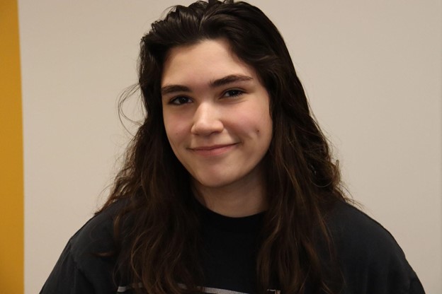 A headshot of Rebecca. Rebecca is wearing a black t-shirt. She has loose wavy dark brown hair. Rebecca is smiling.