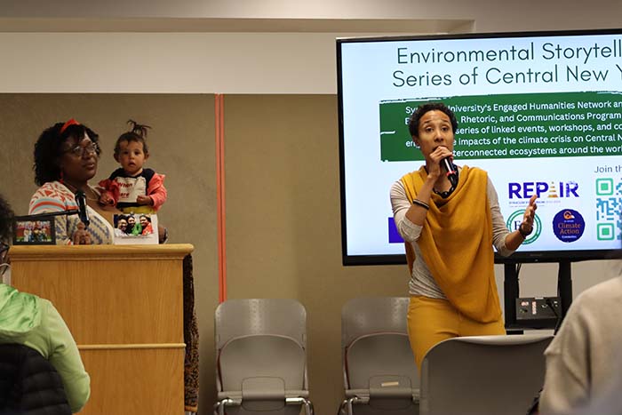 Sarah Nahar, a coordinator for the Environmental Storytelling Series, addresses the audience at an event. She has short hair and is wearing a yellow shawl. Her daughter is held by SeQuoia Kemp in the background.