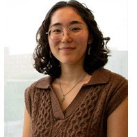A headshot of Shannon Dunn. She is giving a soft smile. Her hair is dark . She is wearing thin-framed glasses and a brown sweater.