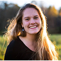 A headshot of Siela Zembsch. She is wearing a black sweater. She has long dirty blonde hair, which is glowing in the sunlight. She is smiling with her teeth. She is standing in a field of yellow flowers.