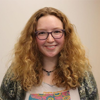 A headshot of Sophia. She is wearing a white t-shirt with a square pink, yellow, and blue design. She has a sweater on. Sophia has blonde curly hair. She is smiling with her teeth. 