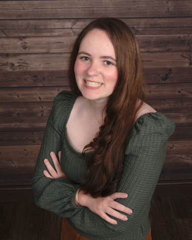 A headshot of Sydney . They are wearing a long sleeved, green, scoop-necked blouse. Sydney is smiling with their teeth.  They have long brown hair. Sydney is standing in front of horizontal dark, wooden panels.