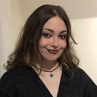 A headshot of V O’Brien. They are wearing a dark blue patterned sweater. They have long brown hair. They are smiling.